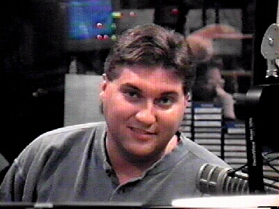A radio DJ smiling while sitting in a studio, with audio equipment and CDs visible in the background.