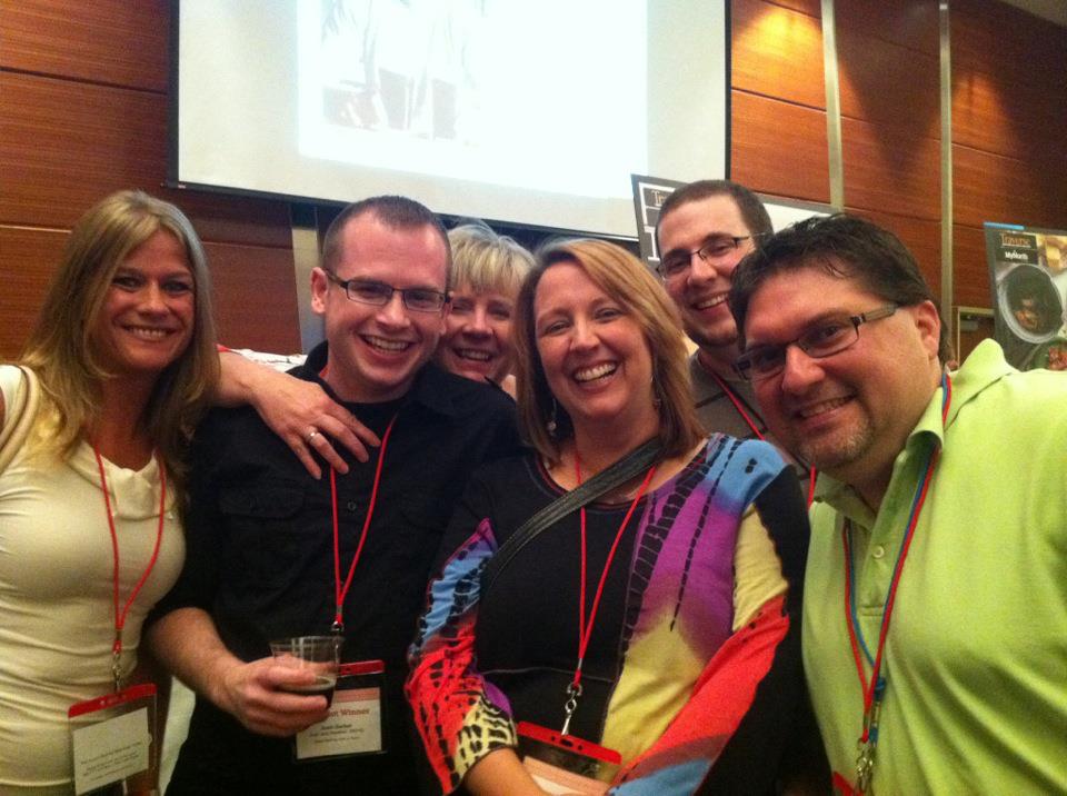 Group of six people smiling and posing for a photo at a social event, wearing name tags and enjoying a festive atmosphere.